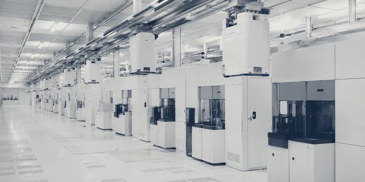 A wide aisle inside a semiconductor fabrication plant: sealed wafer carriers suspended from an overhead transport rail, with a row of enclosed process tools below.